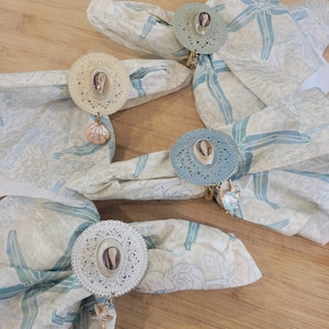 Könnte beinhalten: Vier weiße und blaue Serviettenringe mit Muschelmotiven und Anhängern. Die Ringe befinden sich auf weißen Servietten mit einem blauen Seesternmuster.