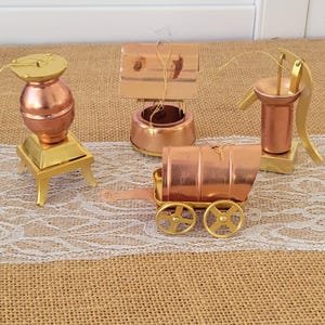 May include: A collection of miniature copper and gold-toned ornaments. The set includes a stove, a well, a water pump, and a wagon. Each ornament has a gold string for hanging. The ornaments are displayed on a burlap and lace surface.