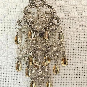 May include: Silver brooch with a crown design and elaborate filigree. The brooch has gold-coloured, teardrop-shaped dangles, creating a layered, ornate look. The background is a white, textured fabric.
