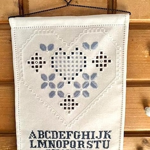 Op de afbeelding: Witte stoffen wandhanger met een hartvormig kruissteekontwerp in blauw en bruin. Het ontwerp bevat bloemen- en geometrische patronen. Het alfabet is in blauw geborduurd aan de onderkant. Het hangt aan een decoratieve metalen hanger.