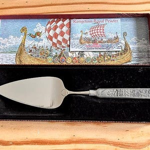Pode incluir: Uma espátula de bolo prateada com uma alça decorativa, apresentada numa caixa com uma ilustração de um navio viking. A espátula tem uma borda serrilhada e é feita de estanho.