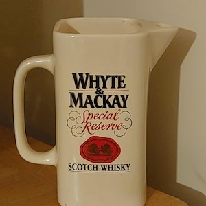 May include: Cream-colored ceramic pitcher with a handle and spout. The front features the text "WHYTE & MACKAY Special Reserve SCOTCH WHISKY" in black and red lettering, with a red seal emblem. The pitcher is rectangular with rounded edges.