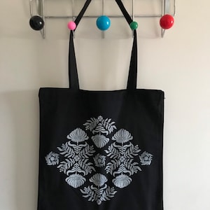 May include: A black tote bag with a white floral design. The bag has two black straps and is hanging on a rack with colourful balls.