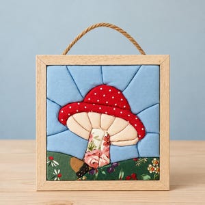 May include: A square fabric wall hanging in a light wood frame with a rope hanger. The textile art features a pieced quilt design of a red mushroom with white dots, a cream-colored underside, and a floral stem, set against a light blue sky.