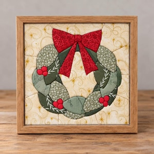 May include: A square framed fabric wall art piece featuring a quilted wreath design. The wreath is composed of various sage green patterned fabric segments accented with bright red berries and a large red bow, set against a cream background.