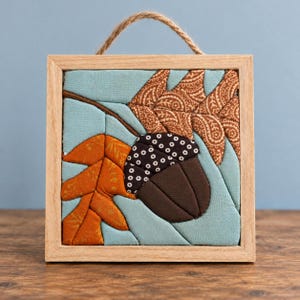 May include: A square fabric wall hanging in a light wood frame with a jute hanger. The textile art features a quilted design of a brown acorn with a black and white polka dot cap, flanked by an orange leaf and a brown paisley leaf on a light blue background.