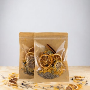 May include: Two stand-up pouches made of brown kraft paper with clear plastic windows. Inside are dried orange slices, cinnamon sticks, yellow flower petals, and purple lavender buds. Loose dried ingredients are scattered in front of the pouches.