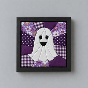May include: A square framed fabric art piece featuring a central white ghost applique with black eyes and a mouth. The background is composed of various purple and white fabric patches in floral, polka dot, and gingham patterns within a black frame.
