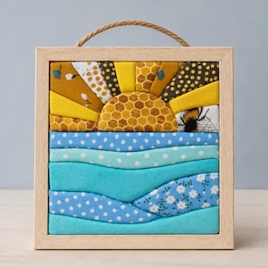 May include: A square fabric wall art piece in a light wood frame with a hanging loop. The top half features a sun with yellow, gold, and honeycomb-patterned fabric rays. The bottom half depicts wavy blue fabric layers with white dots and floral prints.