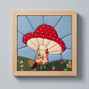 May include: A square framed fabric art piece featuring a pieced quilt design of a red mushroom with white polka dots. The mushroom has a cream-colored underside and a floral patterned stem, set against a light blue sky and a dark green floral ground.