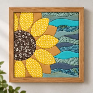 May include: A square fabric wall art piece in a light wood frame featuring a quilted sunflower design. The center is dark brown with white floral patterns, surrounded by yellow polka-dot petals and wavy blue fabric strips representing water.