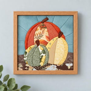 May include: A square fabric wall art piece in a light wood frame featuring a quilted pumpkin patch design. Three fabric pumpkins in orange, sage green, and yellow patterns sit against a blue background with a brown floral fabric base.