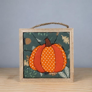 May include: A square wooden frame containing a fabric pumpkin appliqué. The pumpkin is orange with white polka dots and a brown stem, set against a dark green background featuring printed leaves and acorns. A jute twine loop is attached to the top.
