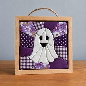 May include: A square wooden frame containing a fabric quilt block featuring a central white ghost with black eyes and mouth. The background consists of various purple and white patterned fabric patches, including floral, polka dot, and gingham prints.