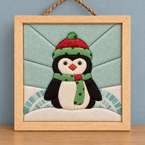 May include: A square fabric wall hanging in a light wood frame featuring a quilted penguin wearing a red hat and a green polka-dot scarf. The background consists of light blue fabric panels arranged in a sunburst pattern above a white snowy base.