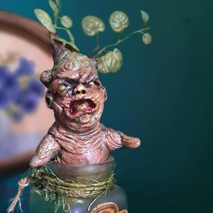 Könnte beinhalten: Ein grünes Glasgefäß mit einem braunen Etikett, auf dem "Alraune Wurzel Salt" steht, und einer kleinen, braunen, humanoiden Figur mit grünen Blättern, die aus ihrem Kopf wachsen. Die Figur befindet sich im Gefäß.