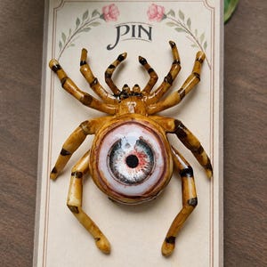 Könnte beinhalten: Eine spinnenförmige Anstecknadel mit einem realistischen, glänzenden, menschlich wirkenden Auge mit blauer Iris und schwarzer Pupille in der Körpermitte. Die Spinne hat acht segmentierte, hellbraune Beine mit dunkelbraunen Streifen auf einer Karte.