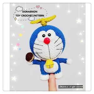 May include: A blue and white crocheted Doraemon toy holding a brown and white pastry. The toy has a yellow propeller on its head and a yellow bell around its neck. The text "Doraemon Toy Crochet Pattern" is written in a white cloud above the toy.