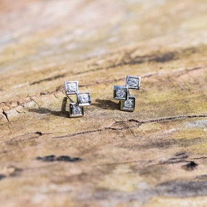 Peut inclure: Une paire de boucles d'oreilles en argent avec de petits diamants de forme carrée. Les boucles d'oreilles sont sur une surface en bois.