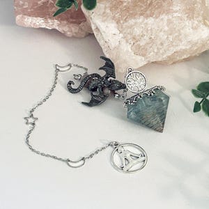 May include: A silver-toned pendulum with a dragon charm, a tree of life charm, and a geometric crystal. The pendulum chain features star and crescent moon charms. The crystal is light blue and faceted.