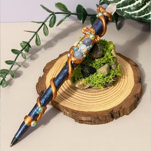 May include: A handcrafted pen with a dark blue, iridescent barrel, decorated with copper-coloured swirls and accents. The pen features light blue and white beads, and a clear crystal at the top. It rests on a wooden slice with moss and small stones.
