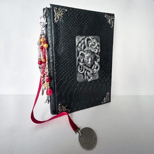 May include: A black journal with a textured snake-skin pattern, featuring silver-toned metal corner protectors and a central relief sculpture of a face with snakes. A silver charm tassel with red and yellow beads hangs from the spine with a red ribbon.