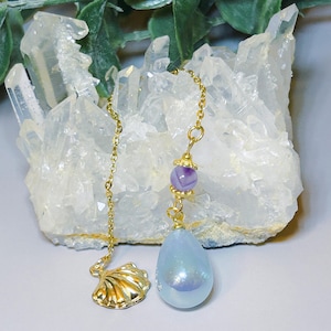 May include: A gold-colored chain with a seashell charm and a light blue teardrop-shaped pendant. The pendant is connected to a gold chain with a purple bead. The seashell charm is on the left side of the image.