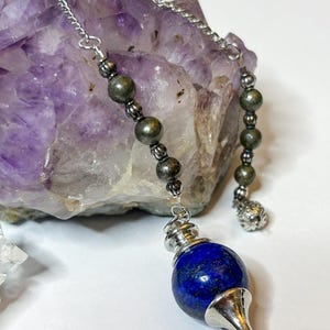 May include: A blue lapis lazuli pendulum with a silver chain and pyrite beads. The pendulum is hanging from a rock.