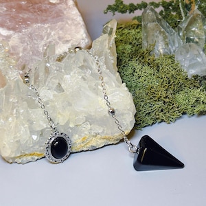 May include: A silver chain pendulum with a black, teardrop-shaped crystal and a round pendant with a black stone. The pendant has a decorative silver border. The background includes quartz crystals and green moss.