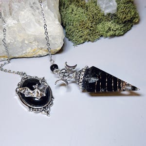 May include: A silver-toned pendulum with a black crystal point and a bat pendant. The pendulum features a crescent moon and star design. The bat pendant is oval-shaped with a silver bat design. The items are on a white surface with a rock and moss in the background.