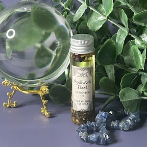 May include: A clear crystal ball on a gold stand, a small bottle of anointing oil labeled "Spellwork Boost", and a blue crystal bracelet are displayed against a backdrop of green foliage. The oil bottle has a silver cap and a decorative label.