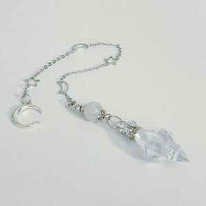 May include: A silver-toned pendulum with a clear crystal point and a chain adorned with crescent moons and stars. A white, round bead is positioned above the crystal. The pendulum is designed for divination and spiritual practices.