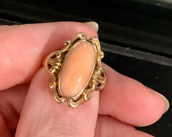 May include: A gold ring featuring a large, oval-shaped coral stone. The coral has a soft, peachy-pink hue, set in a decorative gold filigree setting. The ring is held by a hand.