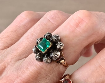 May include: A gold ring featuring a cluster of small, dark stones surrounding a central, emerald-cut green gemstone. The ring has a floral design and is set against a neutral background.