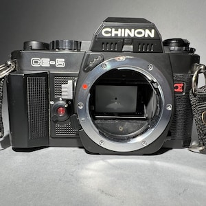 May include: A black Chinon CE-5 35mm film camera with a silver lens ring and a black strap.