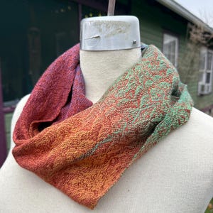 May include: A handwoven infinity scarf in shades of red, orange, and green. The scarf is displayed on a mannequin, showcasing its textured weave and color gradient. The design features a blend of warm and cool tones, creating a unique accessory.
