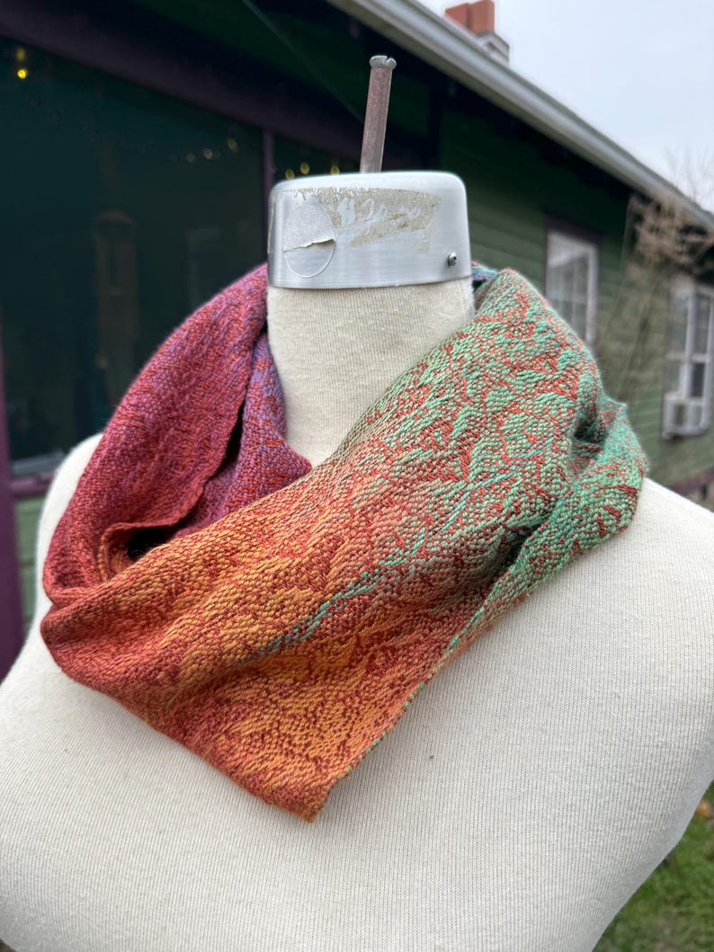 May include: A handwoven infinity scarf in shades of red, orange, and green. The scarf is displayed on a mannequin, showcasing its textured weave and color gradient. The design features a blend of warm and cool tones, creating a unique accessory.