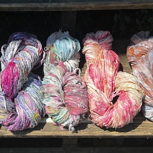 May include: A collection of six skeins of hand-dyed sari silk ribbon yarn, each featuring a unique blend of vibrant colors including pink, purple, blue, orange, and yellow, with a textured, frayed appearance and a soft, lustrous sheen.