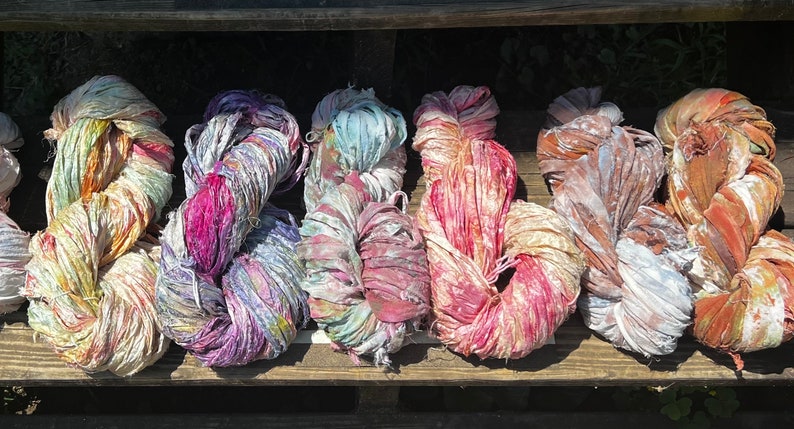 May include: A collection of six skeins of hand-dyed sari silk ribbon yarn, each featuring a unique blend of vibrant colors including pink, purple, blue, orange, and yellow, with a textured, frayed appearance and a soft, lustrous sheen.