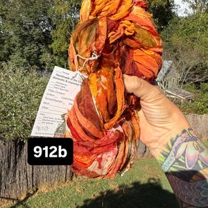 May include: A skein of hand-dyed ribbon yarn in vibrant shades of orange, red, and deep burgundy. A white product tag is attached, featuring the text "Handmade in the US," "Handwoven," "Hand dyed," "Hand Spun Yarn," and the handwritten number "912b."