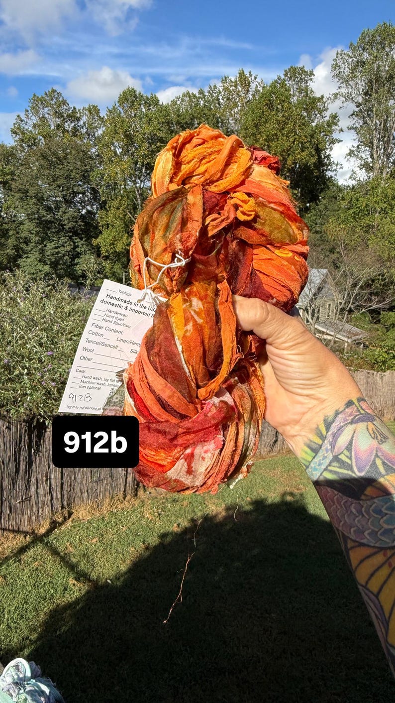 May include: A skein of hand-dyed ribbon yarn in vibrant shades of orange, red, and deep burgundy. A white product tag is attached, featuring the text "Handmade in the US," "Handwoven," "Hand dyed," "Hand Spun Yarn," and the handwritten number "912b."