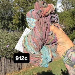 May include: A skein of variegated ribbon yarn featuring a blend of muted red, purple, blue, and teal tones. The yarn has a textured, frayed appearance and includes a white product tag with the text 912c printed in a bold black box.