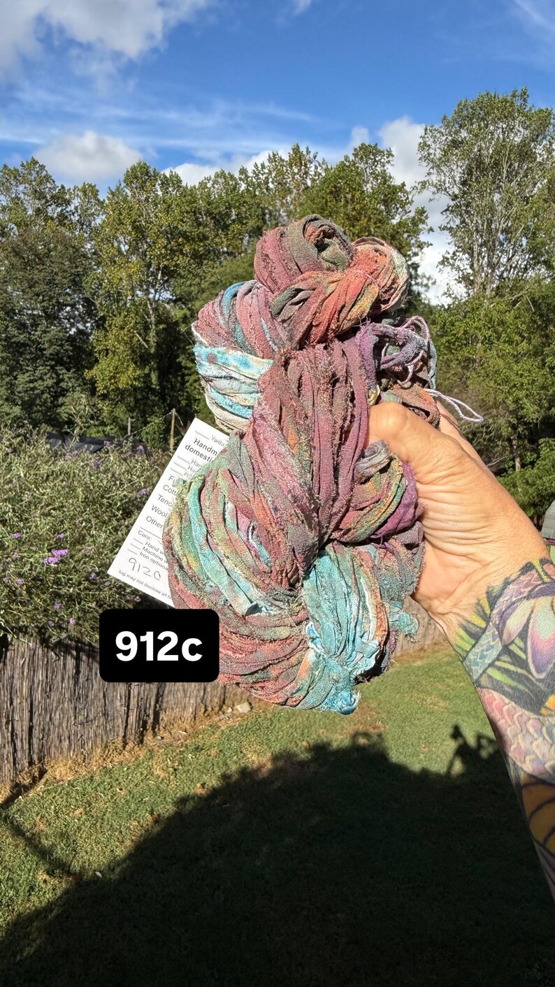 May include: A skein of variegated ribbon yarn featuring a blend of muted red, purple, blue, and teal tones. The yarn has a textured, frayed appearance and includes a white product tag with the text 912c printed in a bold black box.