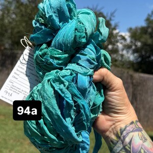 May include: A bundle of teal and turquoise hand-dyed ribbon yarn with frayed edges. A white tag is attached, featuring the text 94a and care instructions including hand wash, lay flat to dry, and machine wash, tumble dry, iron optional.