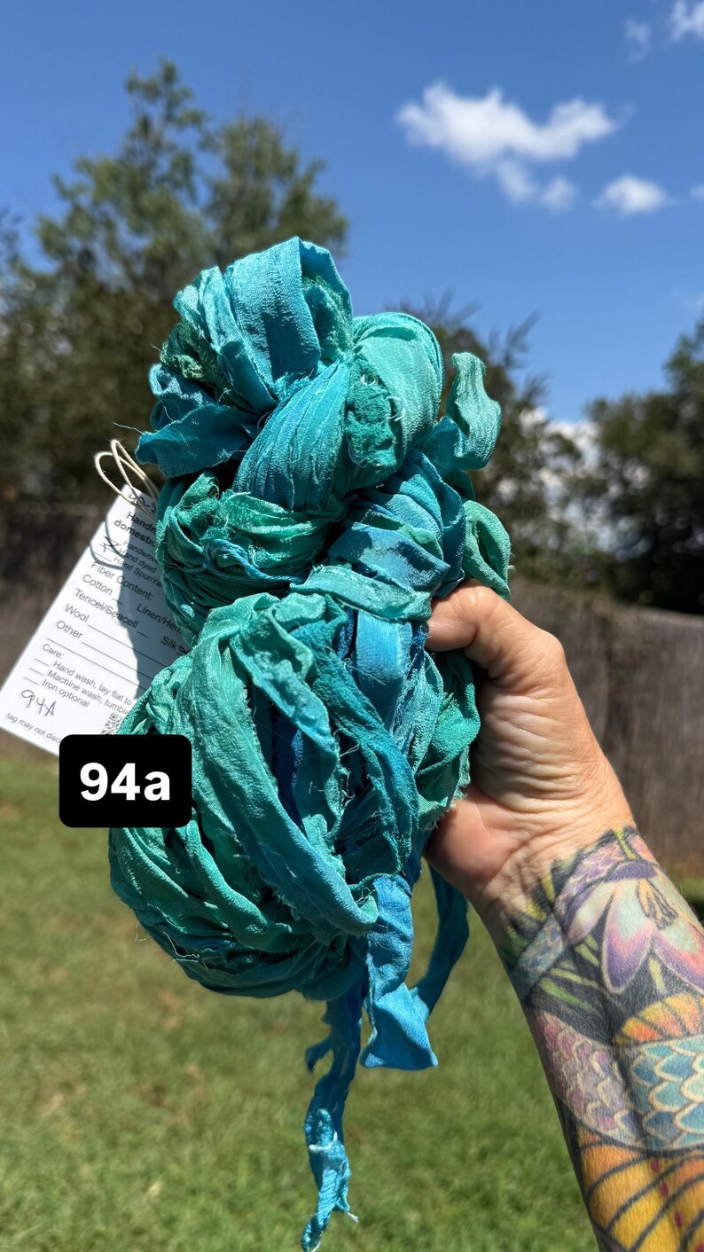 May include: A bundle of teal and turquoise hand-dyed ribbon yarn with frayed edges. A white tag is attached, featuring the text 94a and care instructions including hand wash, lay flat to dry, and machine wash, tumble dry, iron optional.
