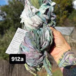 May include: A skein of hand-dyed ribbon yarn featuring a variegated color palette of sage green, soft lavender, and white. A white paper tag is attached, displaying the text 912a and listing fiber content including cotton, linen, and wool.