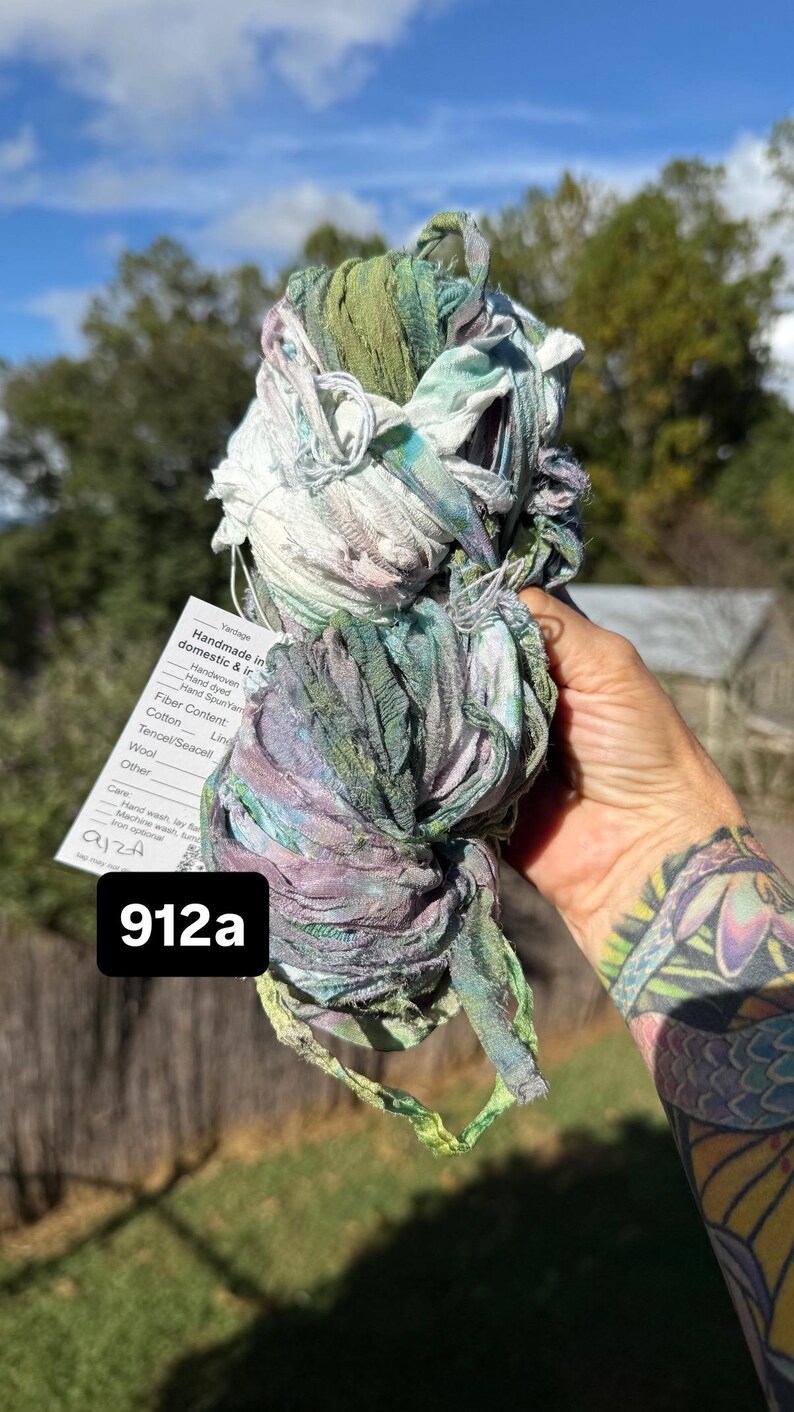 May include: A skein of hand-dyed ribbon yarn featuring a variegated color palette of sage green, soft lavender, and white. A white paper tag is attached, displaying the text 912a and listing fiber content including cotton, linen, and wool.