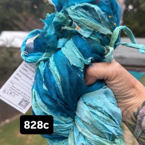 May include: A bundle of hand-dyed silk ribbon in various shades of blue, teal, and turquoise. The ribbon features frayed edges and a crinkled texture. A white product tag is attached, displaying the text 828c and care instructions.