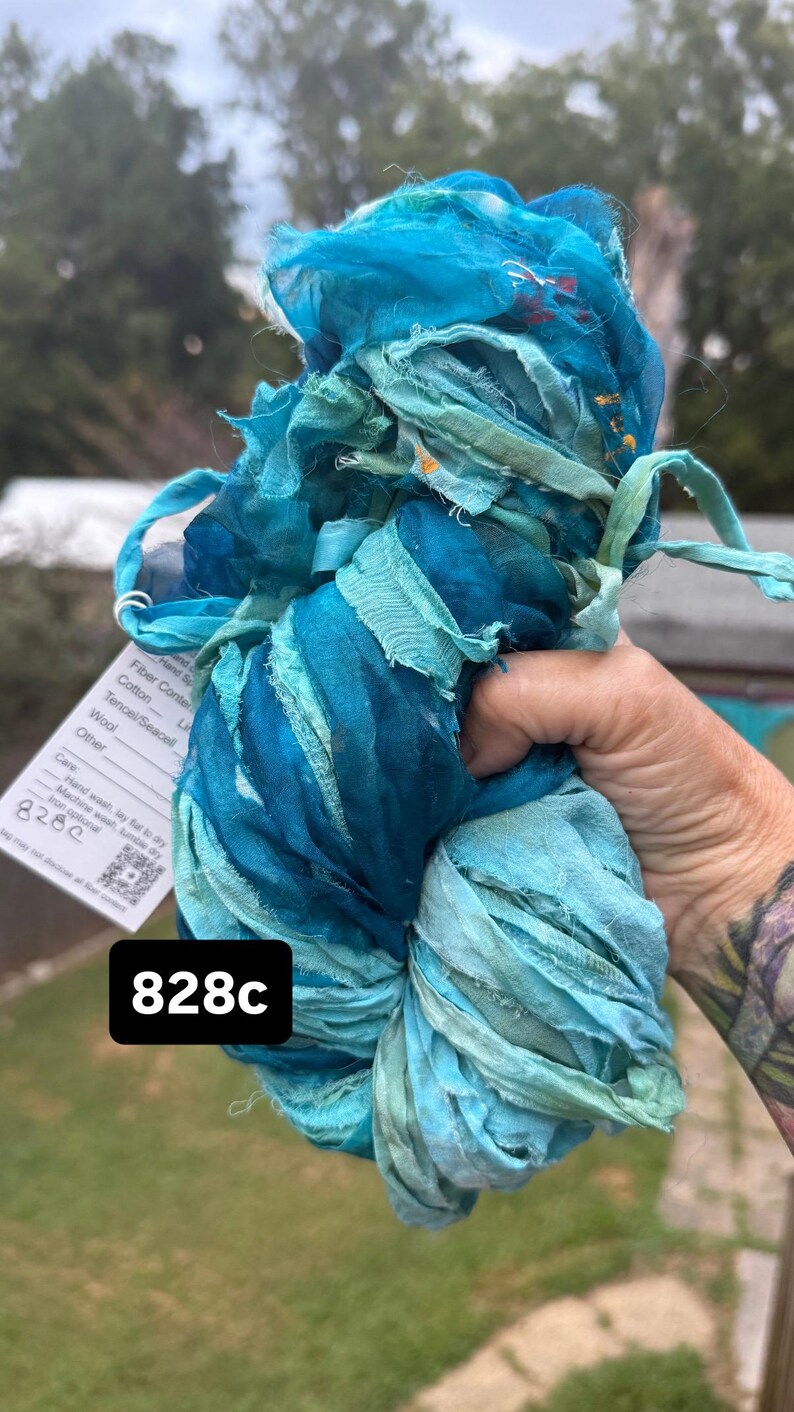 May include: A bundle of hand-dyed silk ribbon in various shades of blue, teal, and turquoise. The ribbon features frayed edges and a crinkled texture. A white product tag is attached, displaying the text 828c and care instructions.