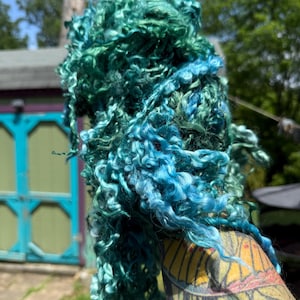 May include: A bundle of curly, long-staple wool locks dyed in a gradient of vibrant teal, emerald green, and bright sky blue. The fibers have a distinct, lustrous sheen and a natural, spiraled texture, showcasing a rich variety of cool tones.